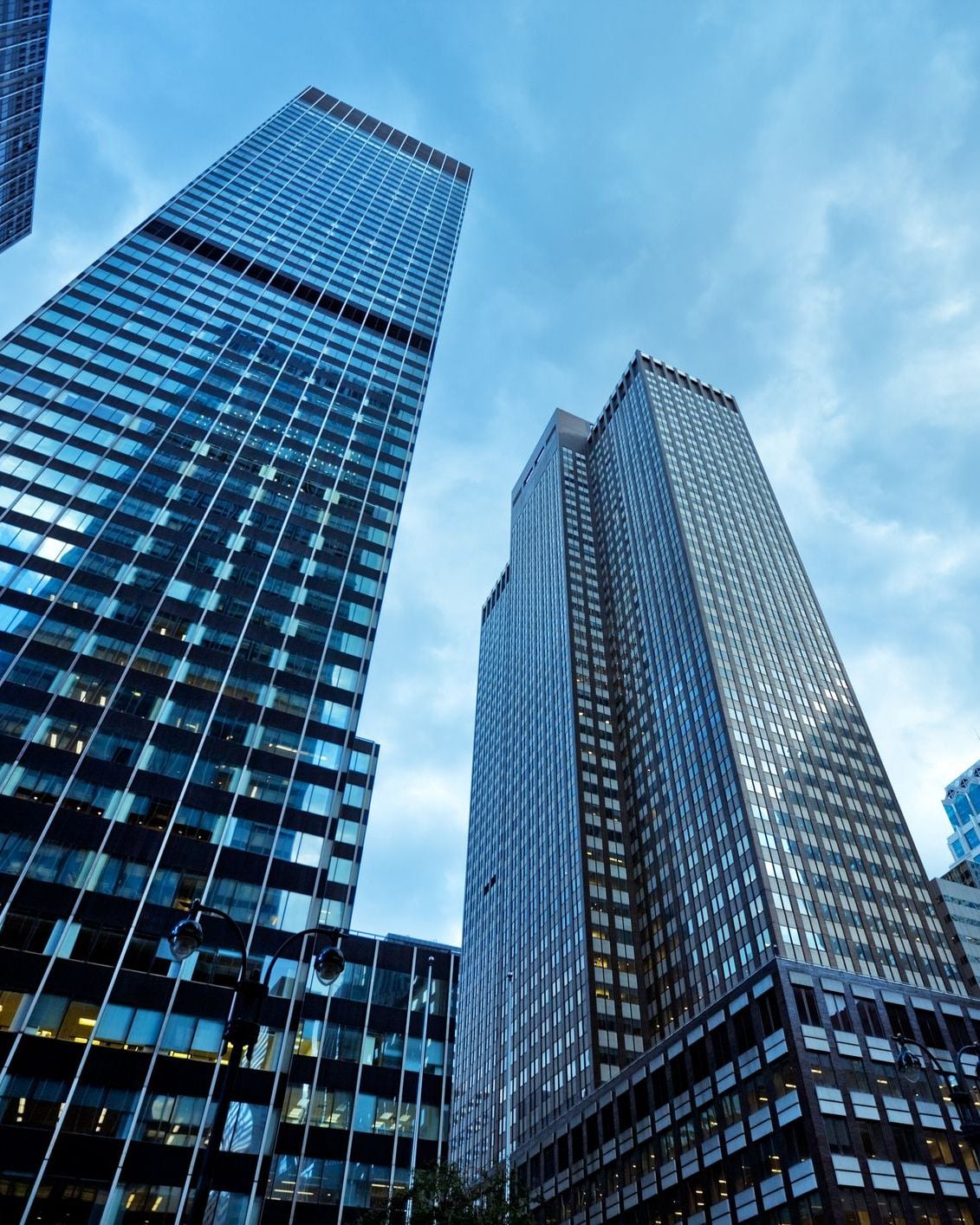Low-angle view of modern glass skyscrapers reaching into a blue sky with clouds, featuring a street lamp in foreground.
