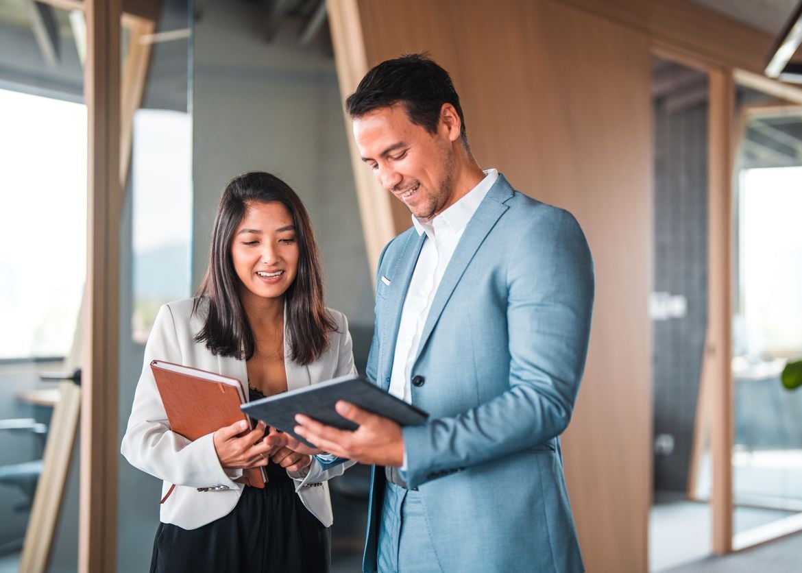 A smiling man in a blue suit and a woman in a white blazer discuss a tablet and notebook in a modern office setting.