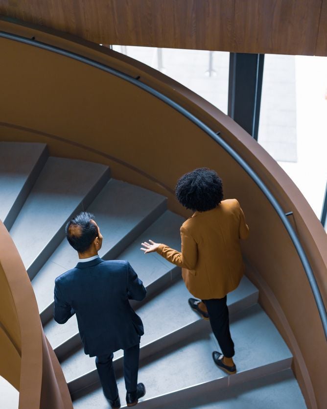 Two professionalsin business attire ascend a modern curved staircase viewed from above.