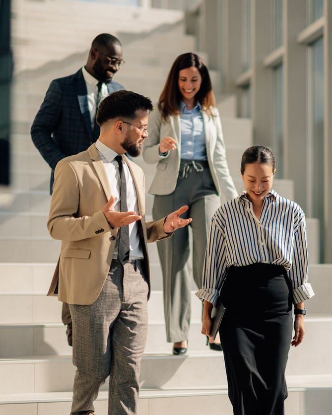 Four business professionals in business attire walking down outdoor steps, smiling and conversing.